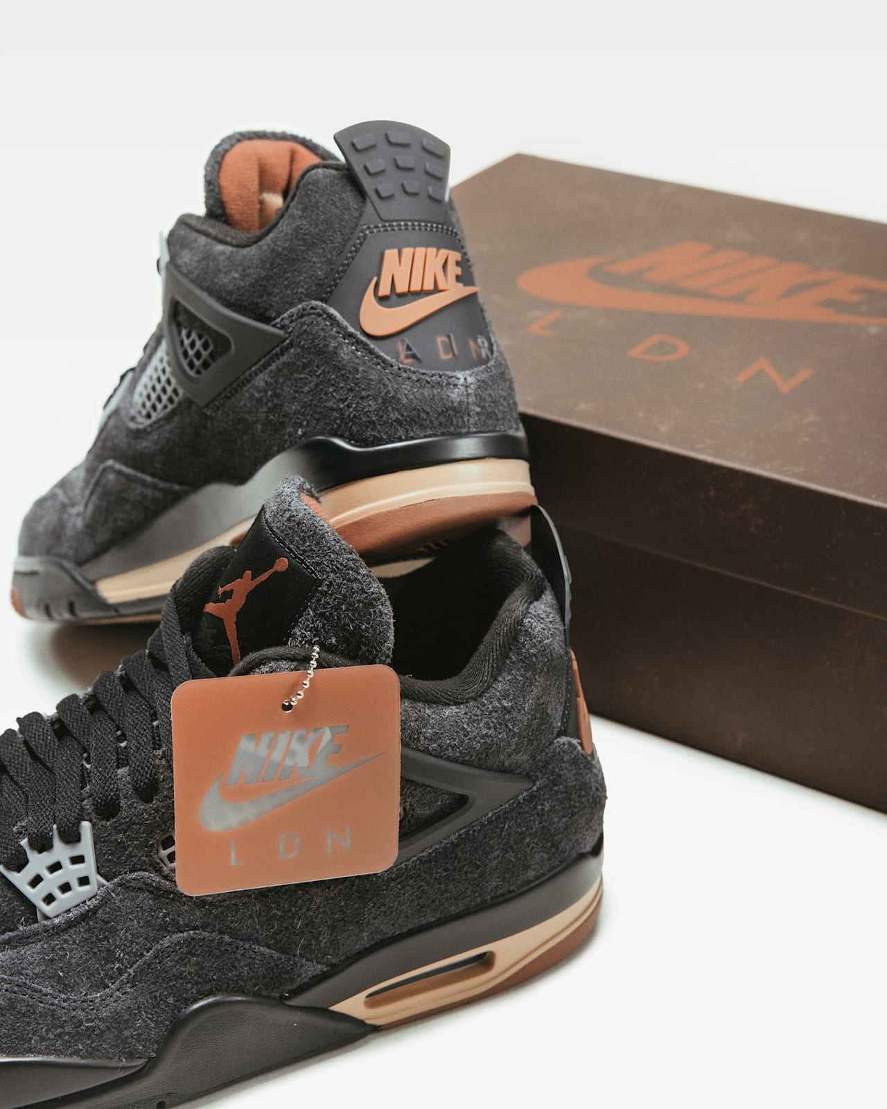 Twee Jordan 4 schoenen, waarbij de ene op de andere steunt op een wit oppervlak met een schoenendoos. Ze hebben een donkergrijs bovenwerk, lichtbruine middenzool en Nike LDN details.