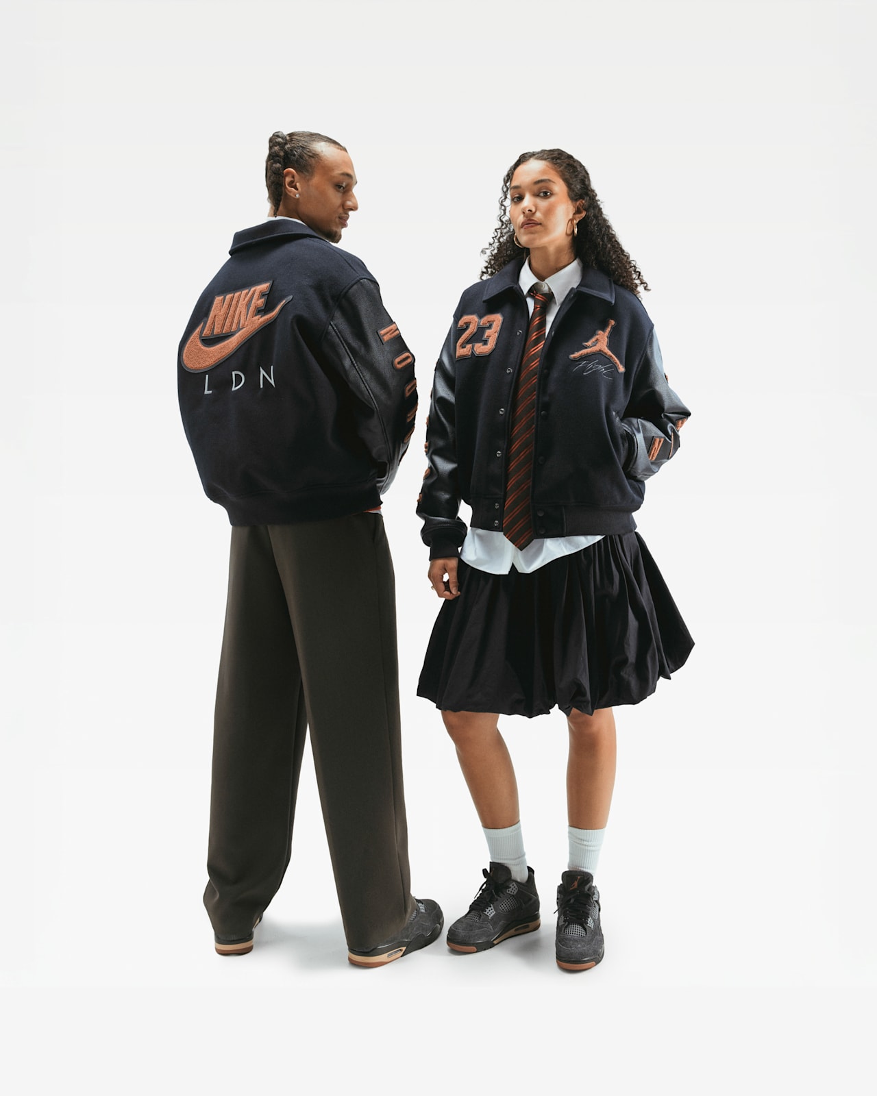 Een man en vrouw staan in een witte studio. Hij draagt een Nike LDN varsityjack en broek. Zij draagt een Jordan varsityjack, shirt, stropdas, rok en Jordan schoenen.