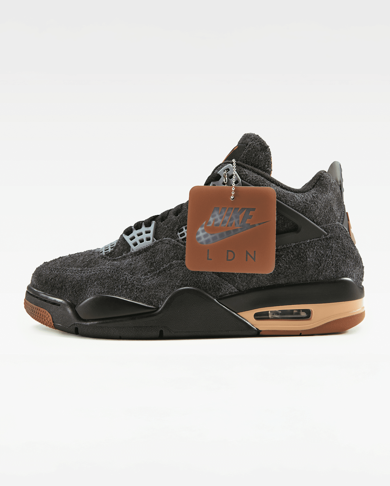 Een Jordan 4 staat rechtop tegen een effen achtergrond. De schoen is donkergrijs met lichtgrijze accenten, een bruine zool en een 'Nike LDN' hanglabel.
