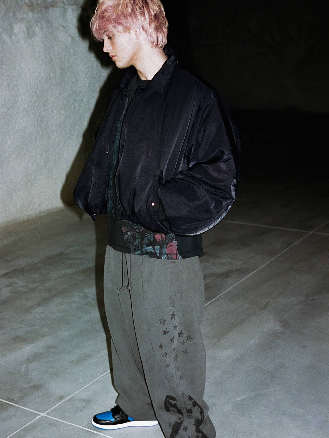 A male model in a black jacket and gray pants stands against a concrete area.