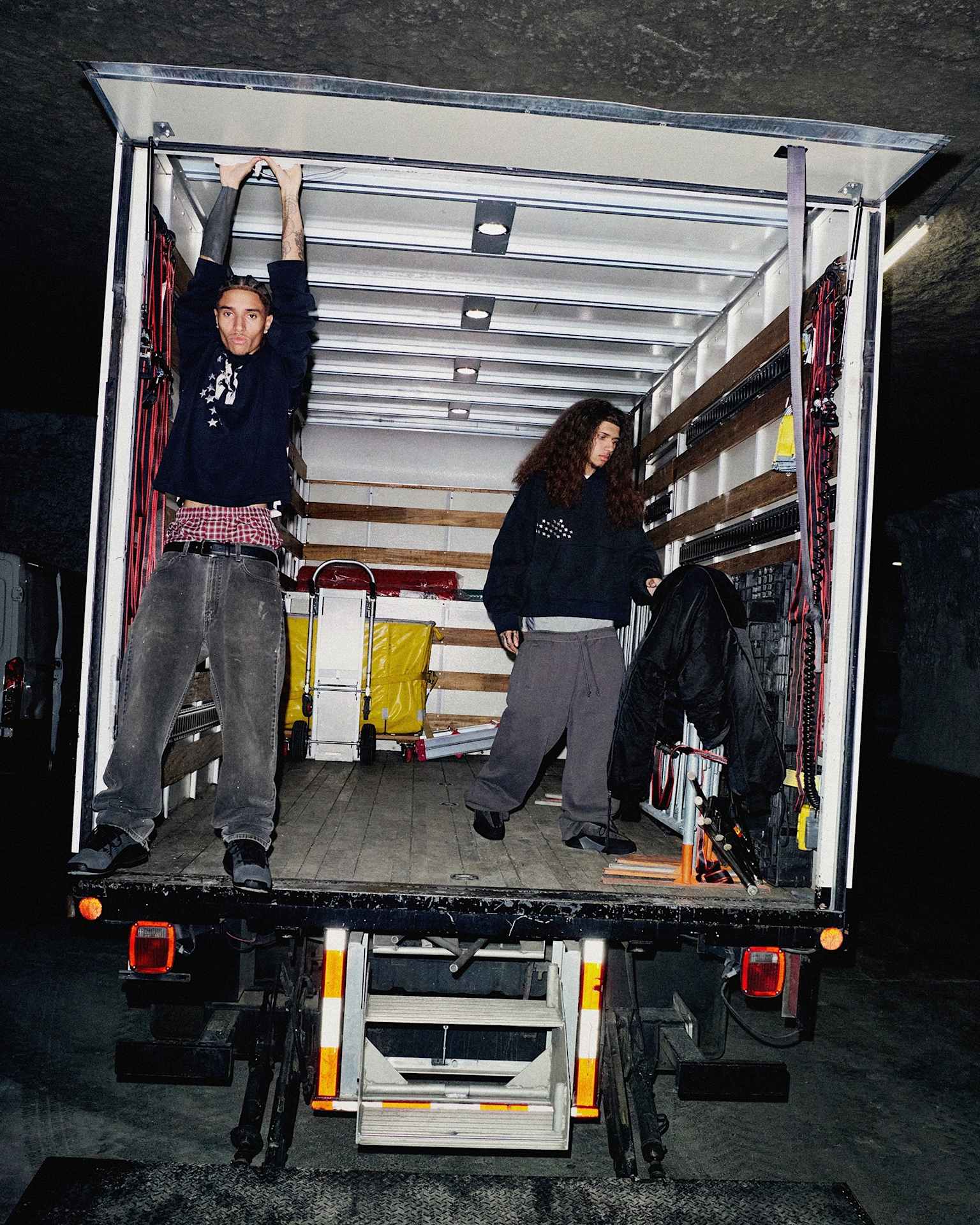 Two male models stand inside an open box truck, filled with equipment and cases.