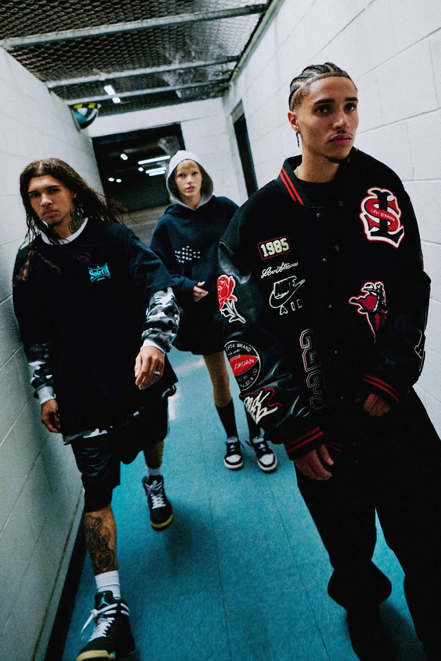 A group of models (two males and one female) wearing black clothing walk through a concrete corridor.
