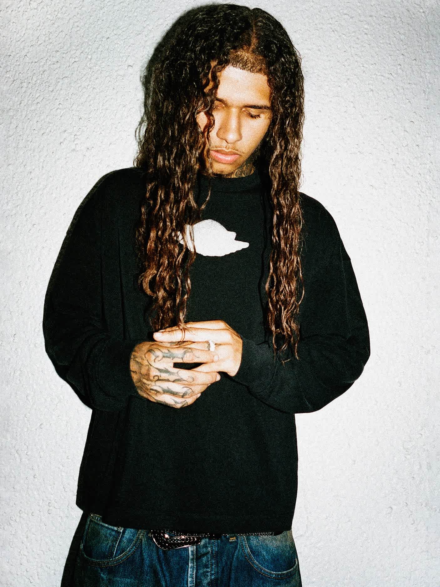 A long-haired male model in a black long sleeved shirt looks down while clasping his hands.