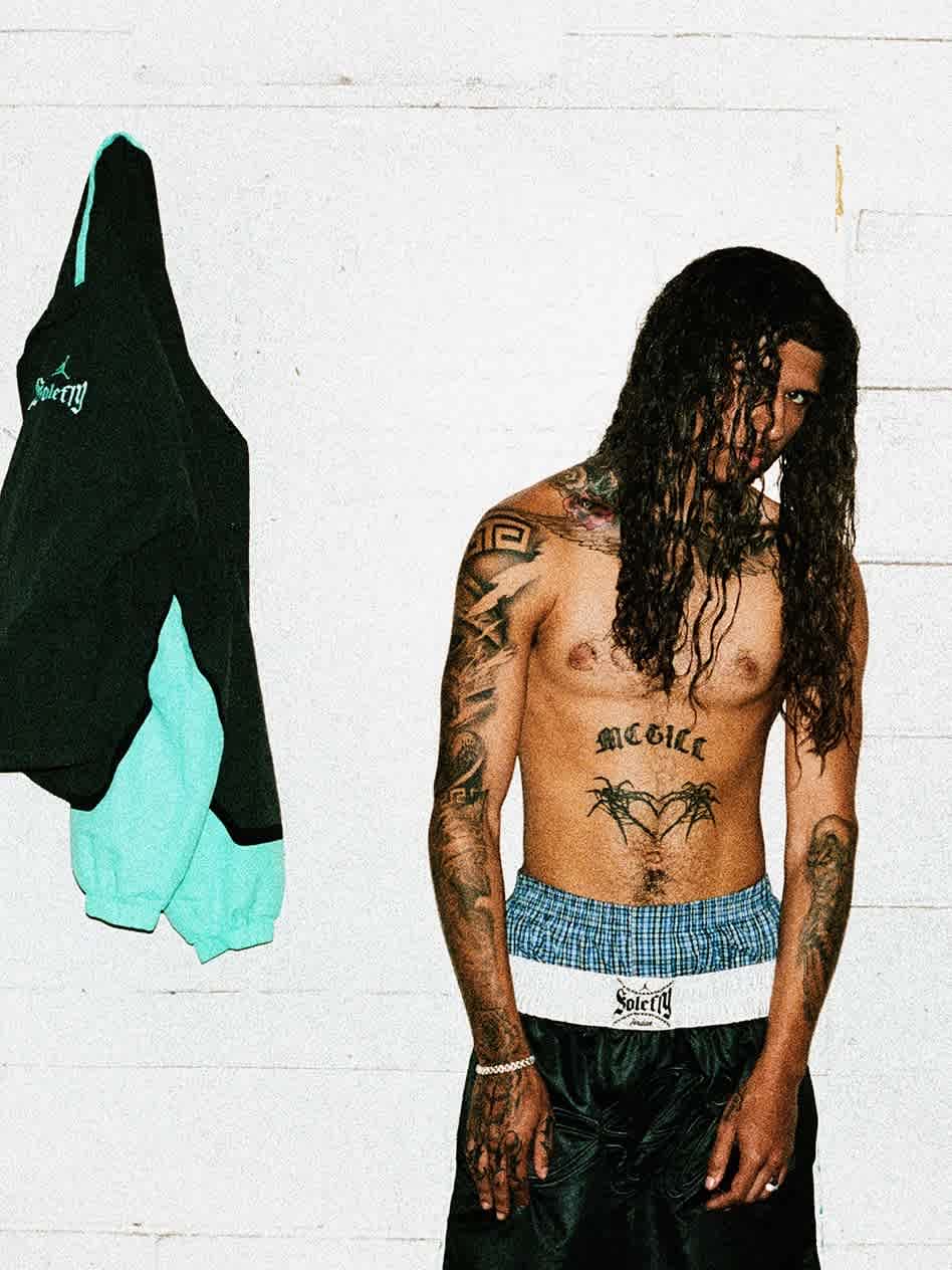 A long-haired male model with tattoos stands in front of apparel hanging on a wall.