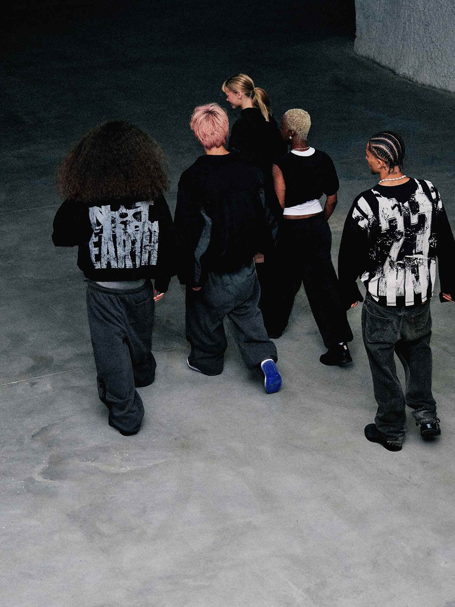 A group of models (three males and one female), dressed in black walk together in a dark industrial space.