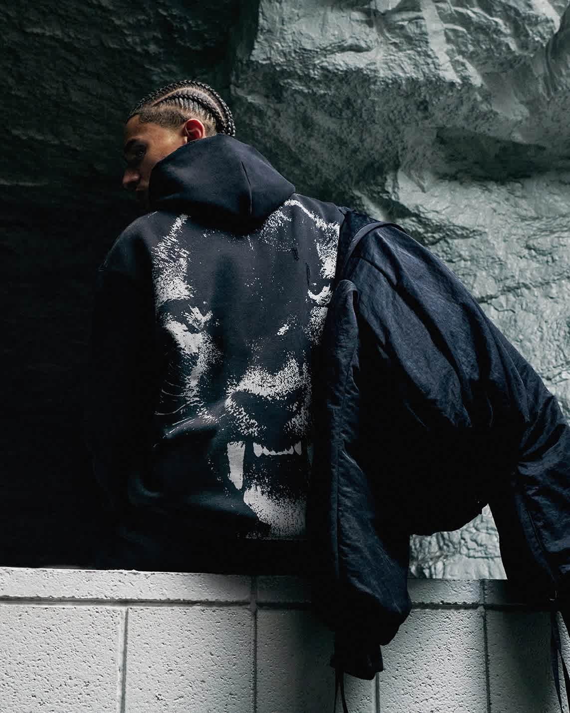 Back view of a model wearing a black hoodie with a panther graphic, and tossing a jacket over their shoulder.