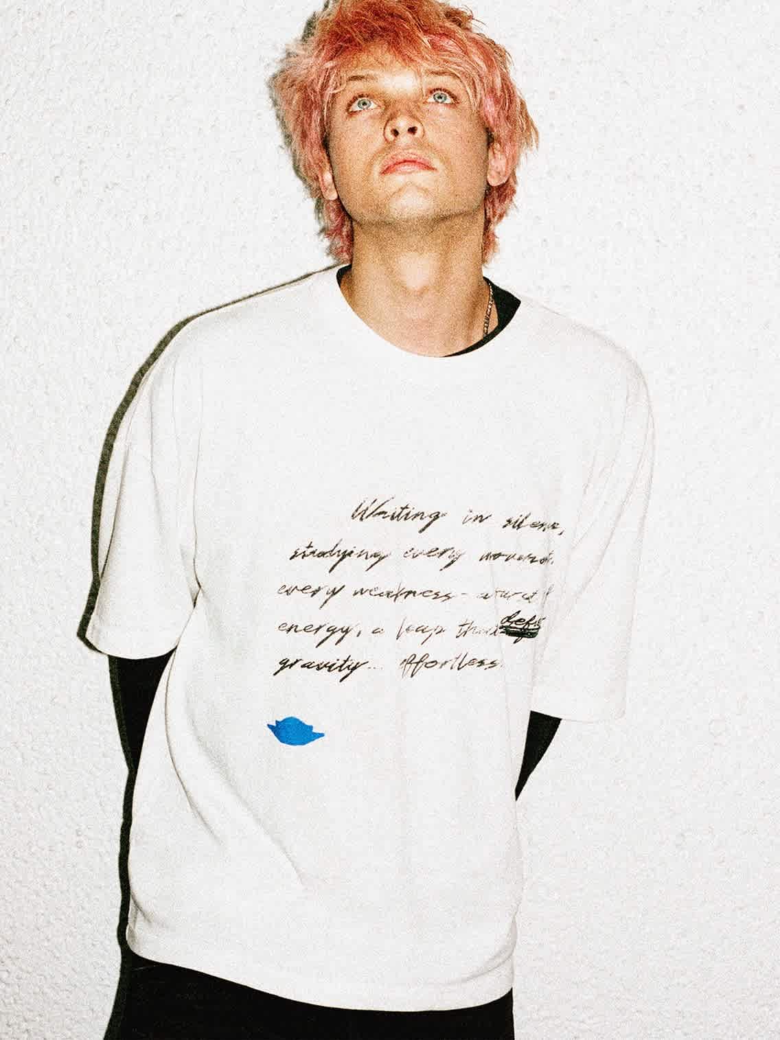 A male model with short pink hair faces upwards in a white long-sleeve shirt.