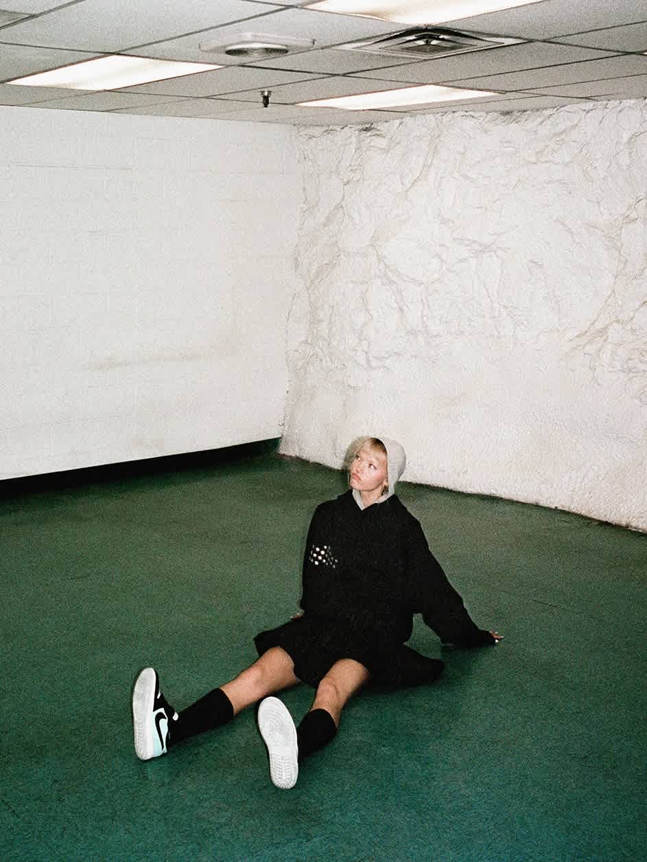 A female model in dark clothing sits on the green tiled floor of an empty concrete space.