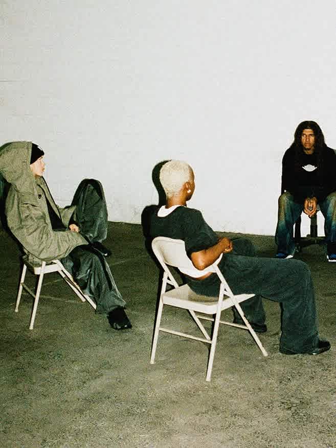 Four models (two females and two males) wearing neutral and dark clothing sit together on chairs in an empty industrial room.