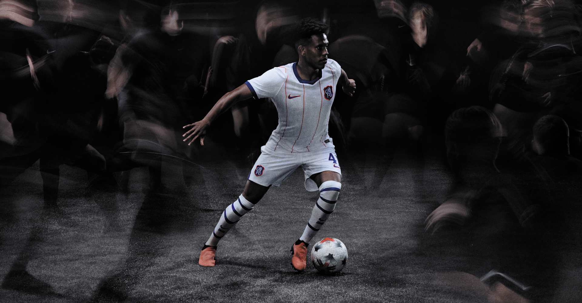 A man dribbling a football on a dark pitch, wearing a white football kit with red and blue accents, and orange football boots.
