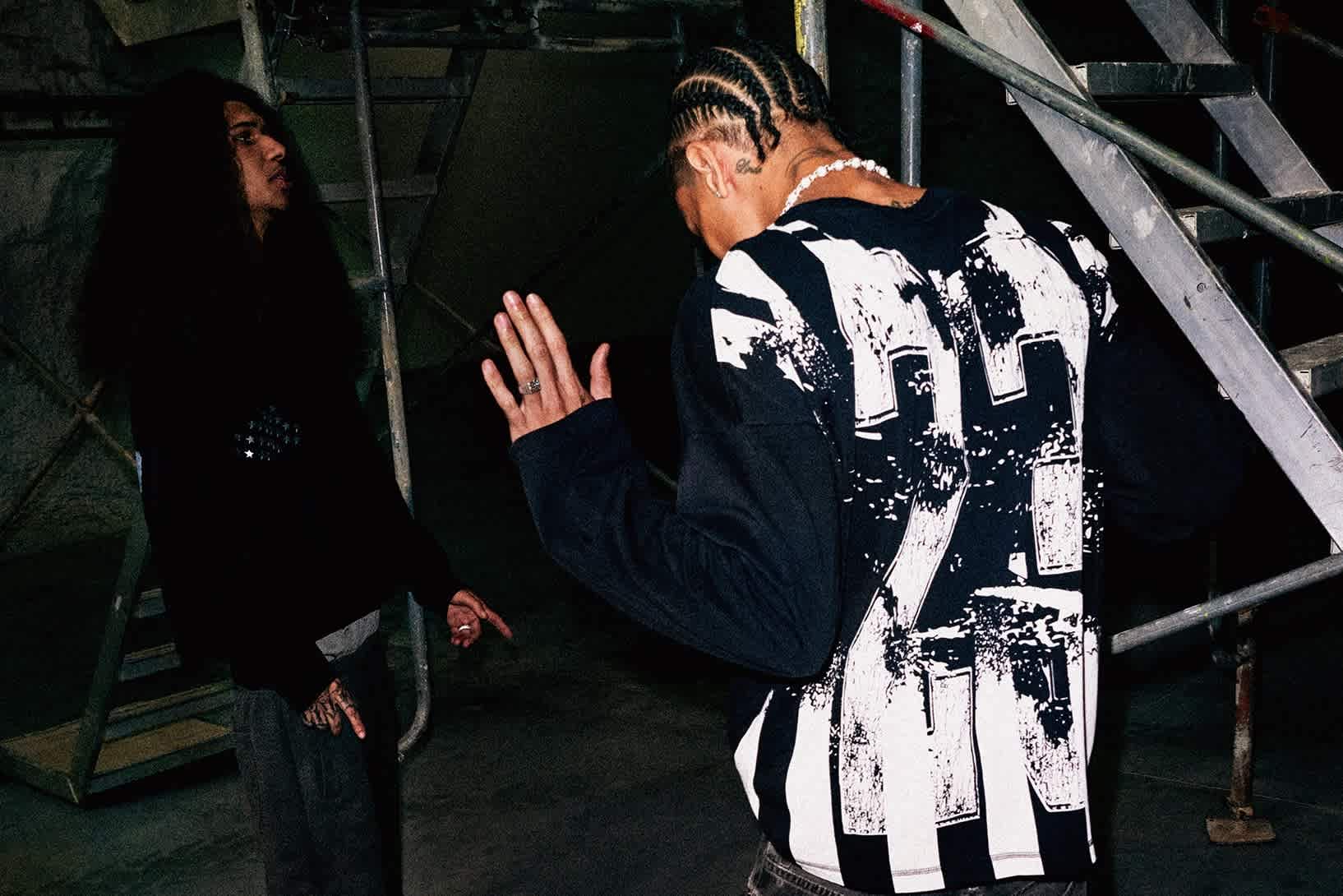 Two male models wearing black long sleeved shirts and black jeans stand in a dark industrial space.