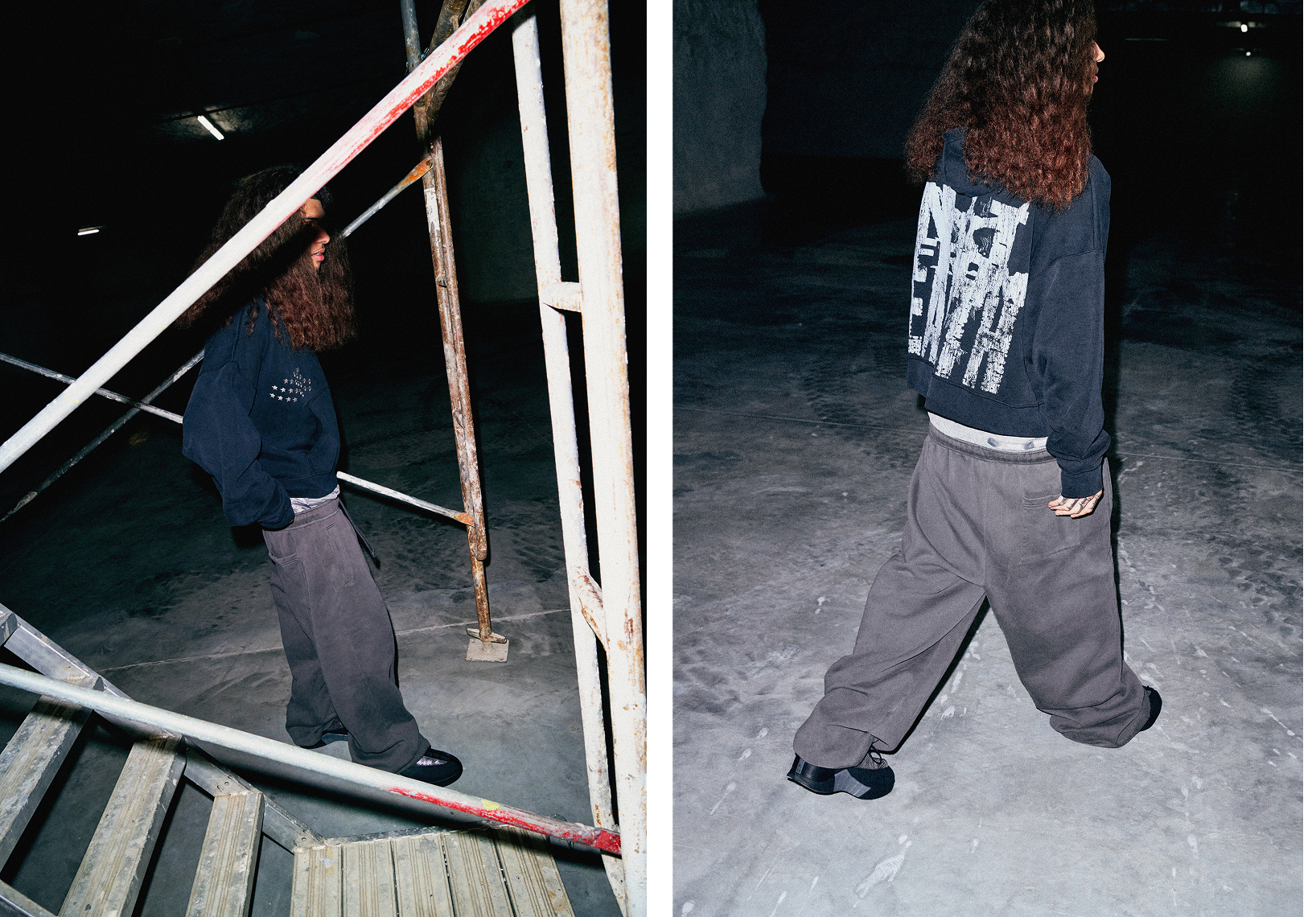 A male model wearing a black long sleeved shirt and black jeans stands in a dark industrial space.