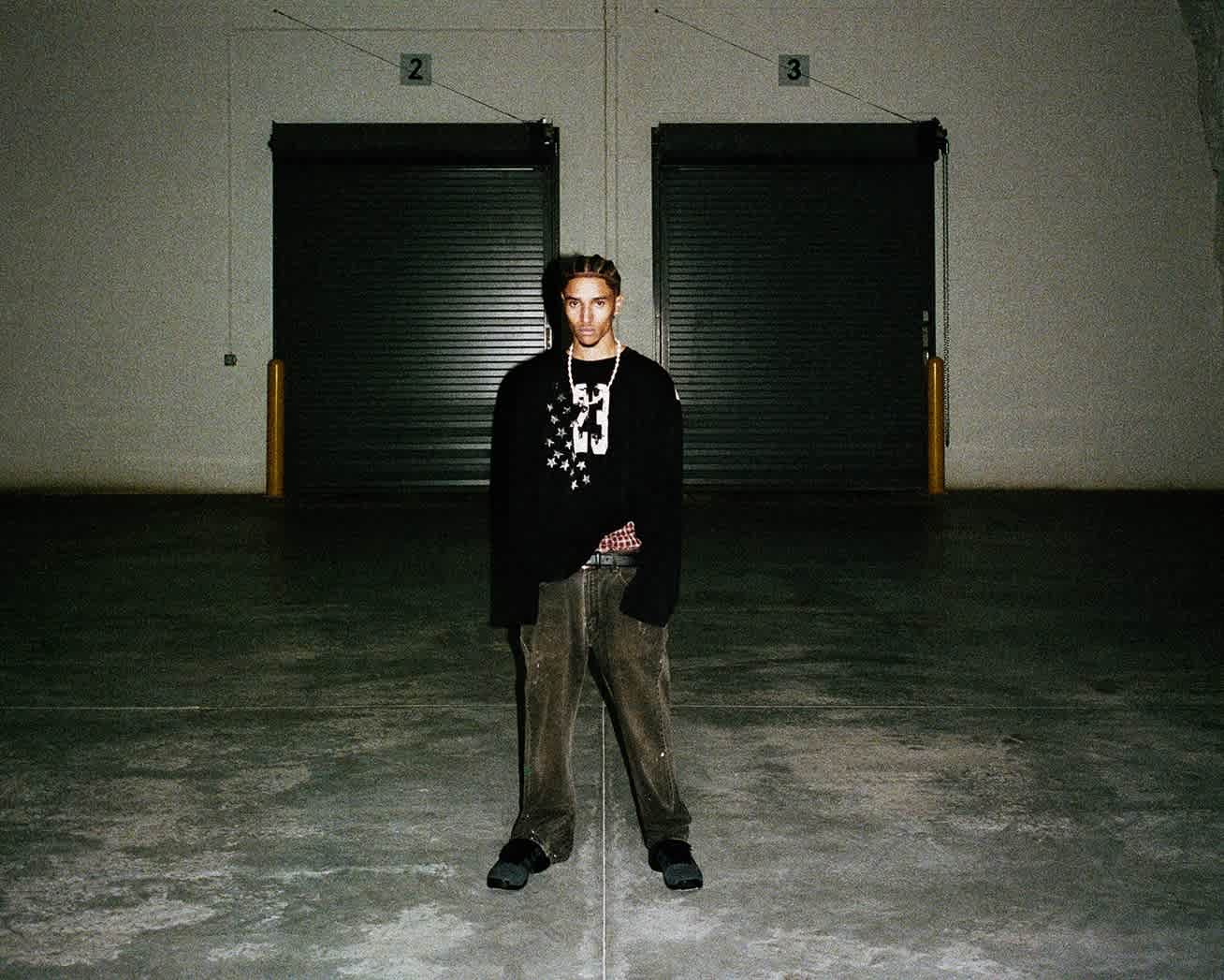 A male model dressed in black stands alone in the center of a large, dimly lit industrial room. He is wearing a black long sleeve with a graphic on the front of it.