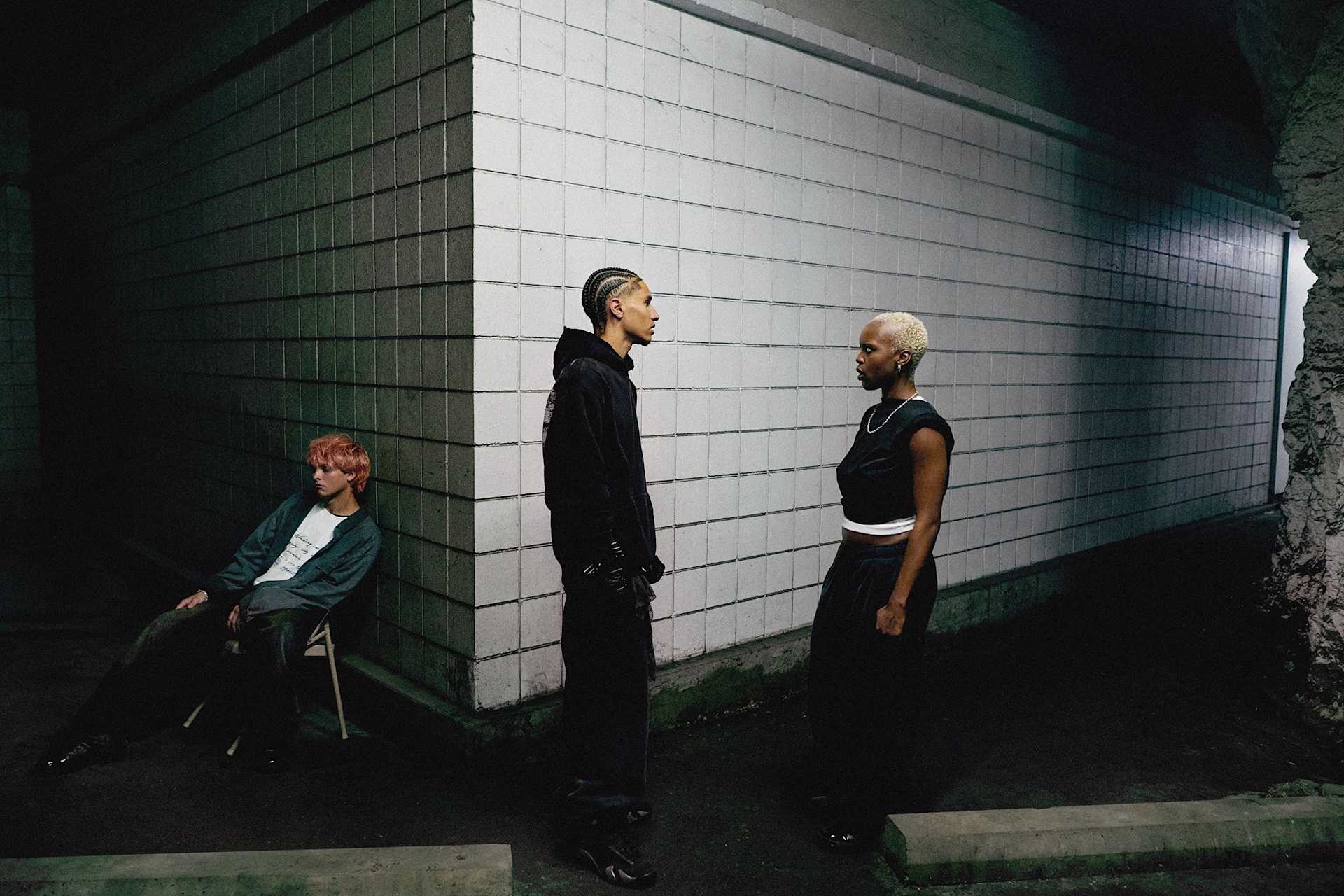 A male and female model in dark clothing stand at different distances in front of a white brick wall. Another male model towards the left sits on a chair in the shadows.