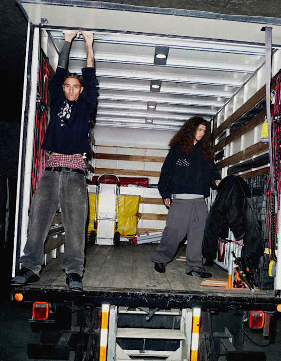Two male models stand inside an open box truck, filled with equipment and cases.